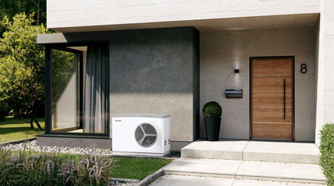 Modern house exterior with a wooden front door and large glass window. A white heat pump unit is placed near the entrance beside a round, green potted plant. The house is surrounded by a well-maintained lawn and small garden.